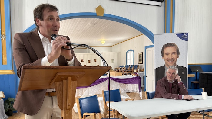 Un homme en veston brun s'exprime au lutrin devant un micro. À sa droite, un autre homme est assis derrière une table blanche, placé devant une bannière de l'Assemblée nationale du Québec illustrant le premier intervenant. 