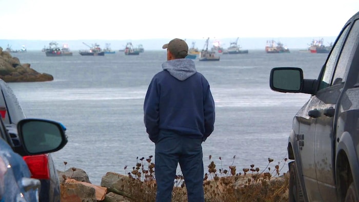 Un homme de dos, sur la berge entre deux véhicules, regarde la mer, où on voit des dizaines de bateaux de pêche dans l'eau.