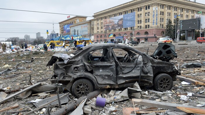 Une voiture carbonisée au centre de la place centrale, entourée de débris de bombardements. 