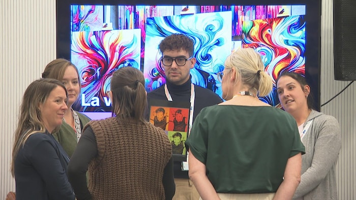 Devant un mur décoré de spirales multicolores, un homme debout tient un carton avec la photo d'un enfant répété quatre fois, mais de couleurs différentes, et cinq femmes l'entourent d'un air confus.