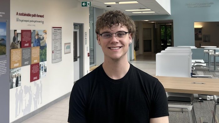 Un jeune homme aux cheveux frisés sourit en posant pour une photo dans un couloir de l'université.