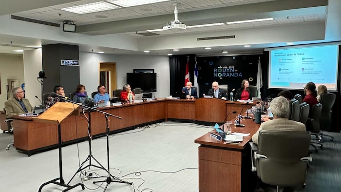 Les conseillers et conseillères municipaux de Rouyn-Noranda regarde un texte afficher sur un toile sur l'effondrement partiel de l'aréna d'Évain.