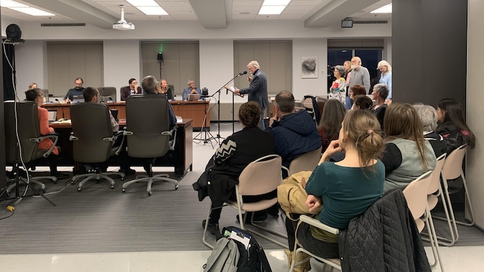 Un homme au micro s'adresse au conseil municipal de Rouyn-Noranda. Derrière lui, des gens debout attendent pour parler à leur tour et d'autres sont assis pour écouter.