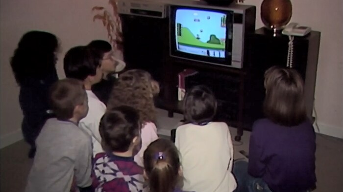 Un groupe d'enfants sont tournés vers un écran de téléviseur sur lequel se déroule une partie du jeu Mario Bros. sur console Nintendo.