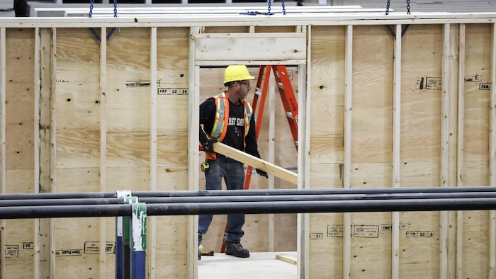 Un travailleur de la construction visible par l'ouverture où une porte sera installée déplace une planche sur un chantier.