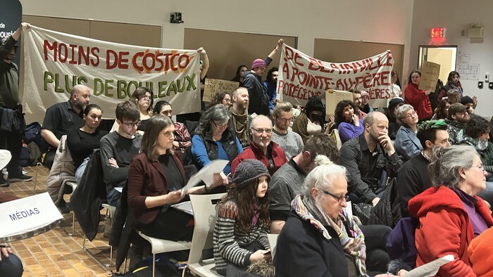 Des citoyens sont assis et d'autres tiennent des banderoles lors de la séance du conseil.