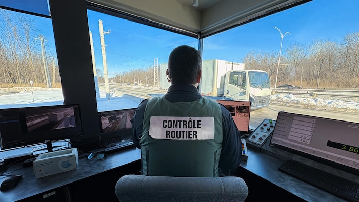 Un contrôleur routier observe un camion lourd par la fenêtre. Il est installé à un bureau, devant un ordinateur.