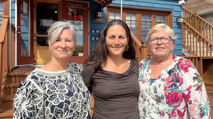 Un portrait de groupe montrant trois femmes souriantes debout sur une terrasse en bois sous le soleil.