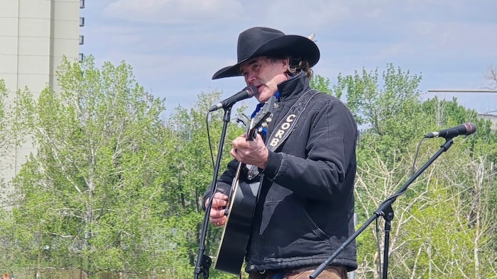 Le chanteur country albertain Corb Lund joue de la guitare acoustique et chante devant un microphone lors d'un rassemblement extérieur.