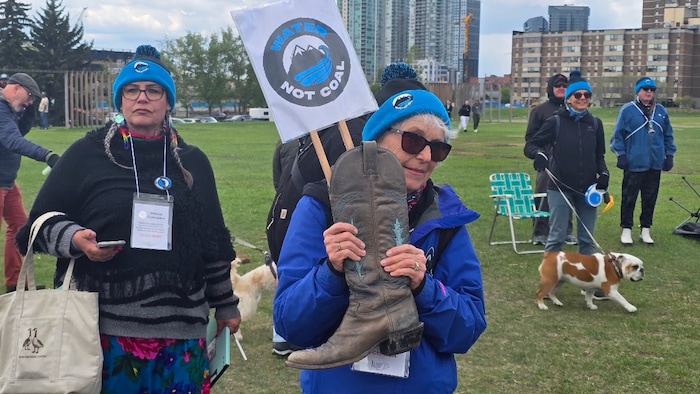 Une femme vêtue d'une tuque bleue et d'un blouson bleu brandit une botte de cowboy en cuir marron surmontée d'une pancarte affichant le slogan « Water Not Coal » (De l'eau, pas du charbon) lors d'un rassemblement.