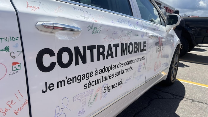 Gros plan sur la portière d'une voiture blanche couverte de signatures manuscrites. Un texte imprimé en noir indique « CONTRAT MOBILE / Je m'engage à adopter des comportements sécuritaires sur la route. Signez ici. » avec le logo de la SAAQ.