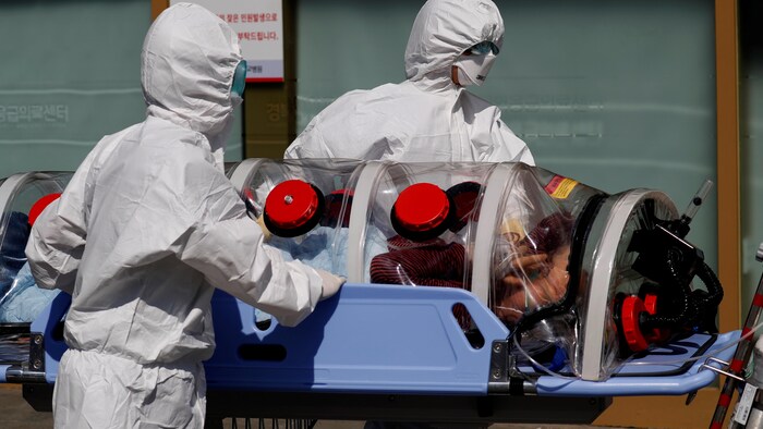 Deux travailleurs portant une combinaison et un masque transportent un patient à l'aide d'une civière d'isolement à pression négative.