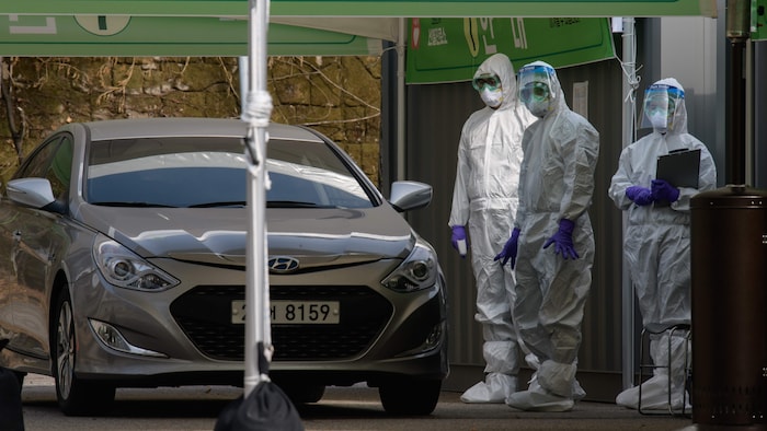 Des travailleurs de la santé portant des combinaisons de protection attendent de contrôler un automobiliste.