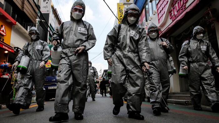 Des soldats portant des combinaisons grises, des gants et des lunettes vaporisent un produit désinfectant dans une rue.