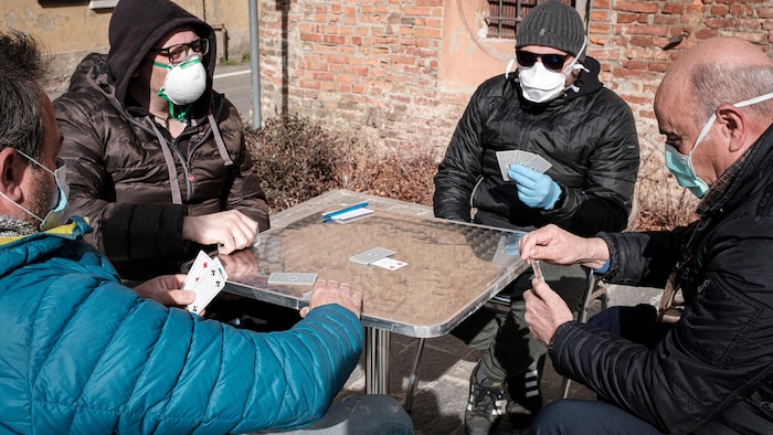 Quatre hommes masqués jouent aux cartes. L'un d'eux porte aussi des gants chirurgicaux.