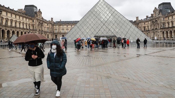 Deux femmes masquées marchent près de la pyramide du Louvre.