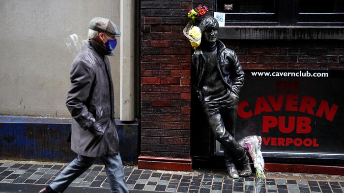 Un passant masqué marchant devant une statue de John Lennon accotée à un pub.