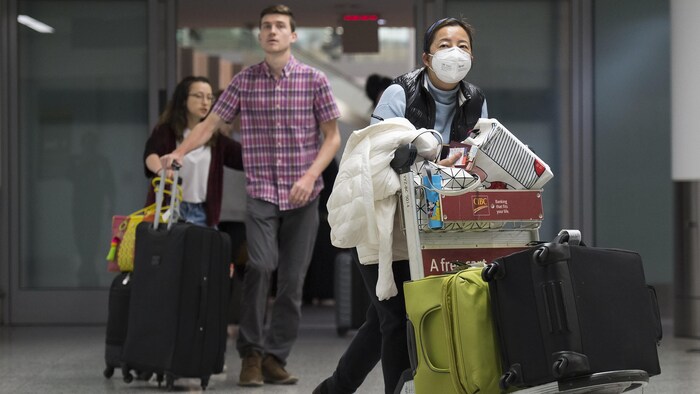 Des voyageurs avec leurs bagages dans un aéroport. Une voyageuse porte un masque.