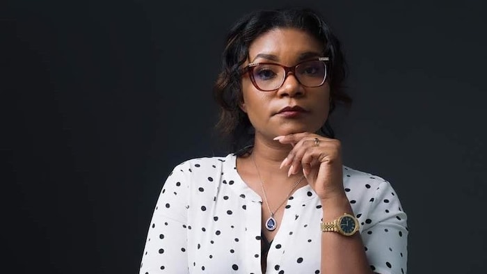 Portrait en studio d'une femme portant des lunettes, un chemisier blanc à pois noirs, un collier avec un pendentif et une montre dorée, qui pose avec la main gauche sous le menton et regarde sérieusement l'objectif devant un fond sombre.