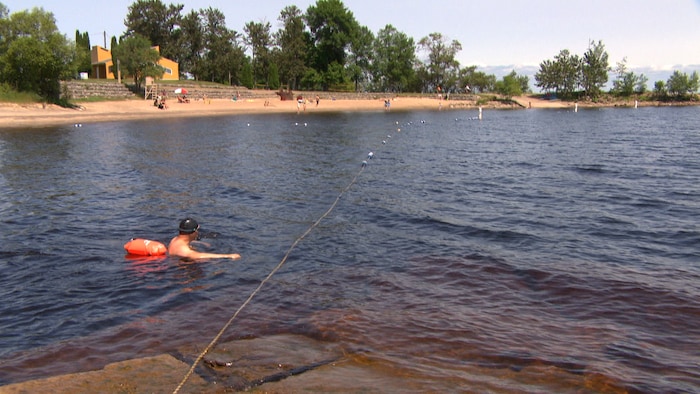 Un nageur portant une bouée est immergé dans l'eau jusqu'à la poitrine. Une plage est visible derrière.