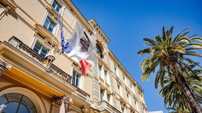 Trois drapeaux flottent sur un bâtiment officiel devant une rangée de palmiers