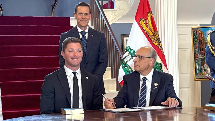 Cory Deagle, souriant, assis à une petite table ronde en bois à côté de Wassim Salamoun, qui le regarde. Le premier ministre est debout derrière.