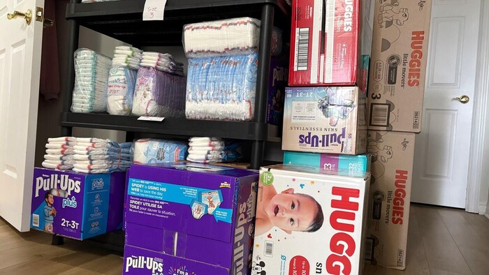 Dans une maison ou un appartement se trouvent des couches pour bébés sur une étagère et des boîtes de couches par terre devant une porte.