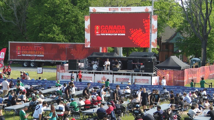 Des affiches rouges dans un parc en plein air et une scène. Des dizaines de personnes prennent place à de longues tables posées sur la pelouse.