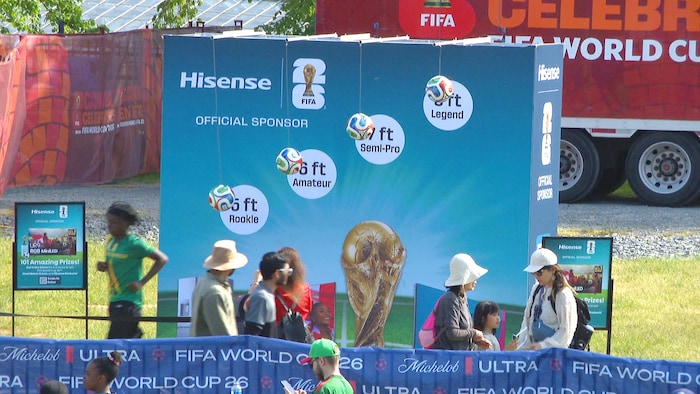 Sur une place publique, un panneau publicitaire avec une photo du trophée de la Coupe du monde et une dizaine de personnes qui circulent devant.