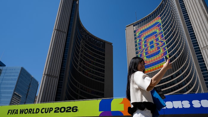 Une jeune femme prend un égoportrait devant le logo de la Coupe du monde de la FIFA 2026 projeté sur l'hôtel de ville, lors d'un événement organisé dans le cadre de la tournée du trophée de la Coupe du monde de la FIFA à la place Nathan Phillips, à Toronto, le lundi 25 mai 2026. 