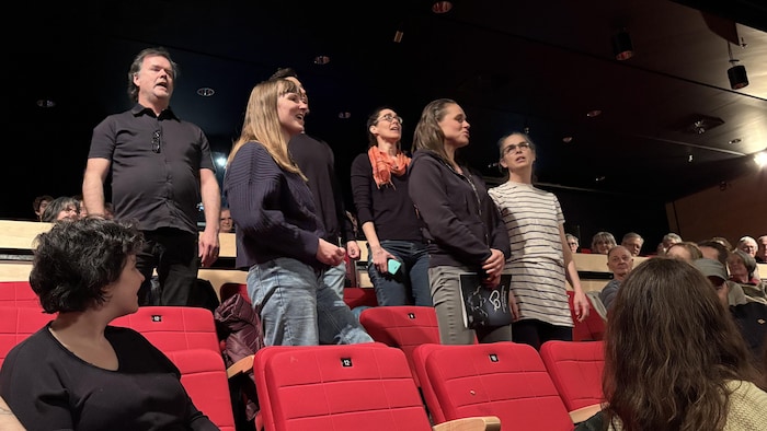 Un groupe de six personne debout dans une salle de cinéma, chantent aux autres spectateurs.