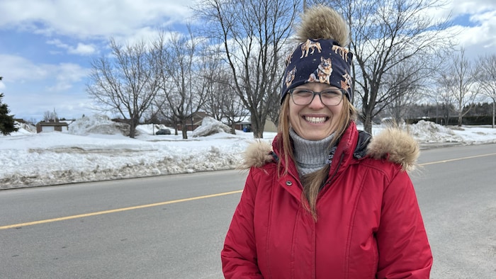 Une femme souriante portant un manteau rouge éclatant et une tuque à pompon pose devant une rue asphaltée. Le paysage en arrière-plan montre des bancs de neige imposants, des arbres sans feuilles et un ciel partiellement nuageux.