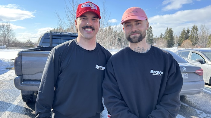 Deux hommes souriants en chandails noirs « Bravo » posent côte à côte dans un stationnement enneigé. Ils portent des casquettes et des gants. Derrière eux se trouvent une camionnette grise, une voiture et des arbres sous un ciel dégagé.