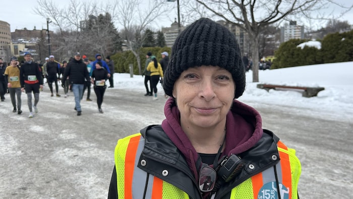 Une femme qui porte une tuque noire et une veste de sécurité jaune fluo sourit au premier plan. Derrière elle, des participants marchent sur un sentier enneigé lors de l'événement sportif « Les 15 km des Pichous » en hiver.