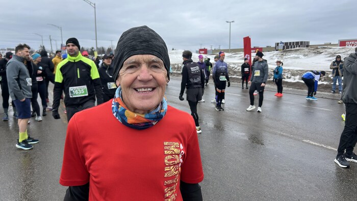 Un homme âgé souriant, vêtu d'un chandail rouge et d'une tuque noire, pose au premier plan. Derrière lui, de nombreux participants se préparent sur une route asphaltée mouillée, bordée de bancs de neige sous un ciel couvert.