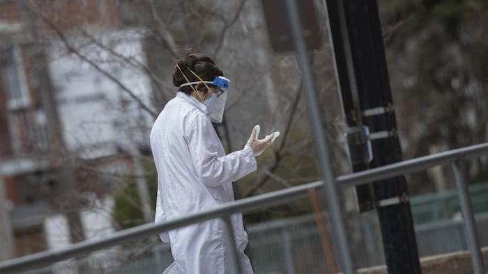 Une femme membre d'une équipe soignante circule à l'extérieur le visage recouvert d'un masque de protection, les mains gantées et vêtue d'un sarrau.