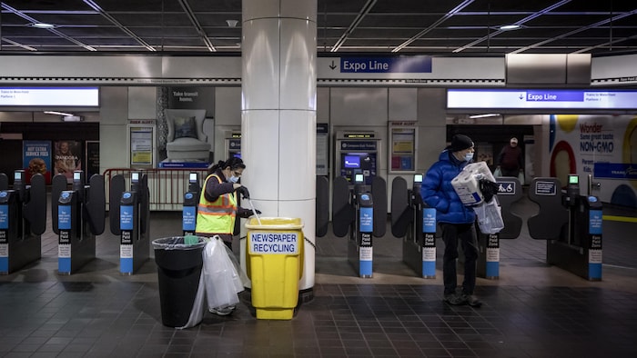 Une travailleuse vide un bac de recyclage alors qu'un homme, qui tient un paquet de papier hygiénique, passe à côté d'elle.
