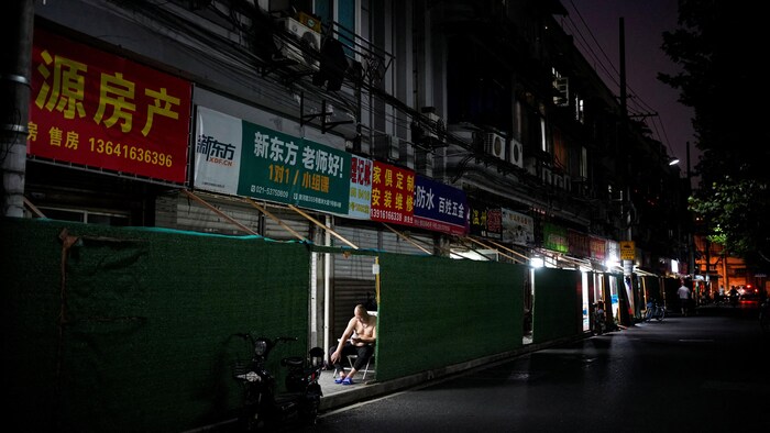 Un homme en bedaine assis sur une chaise derrière une palissade la nuit dans un quartier résidentiel obscur et presque désert à Shanghai.