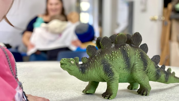 Un dinosaure en avant-plan et une éducatrice et un enfant en arrière-plan.