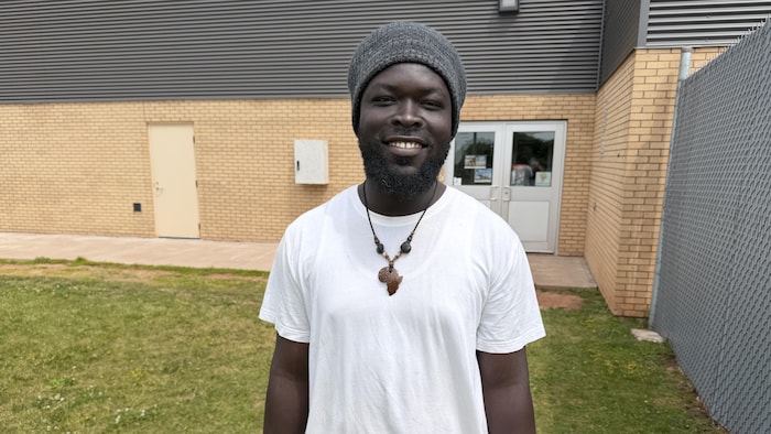 Un homme portant un collier représentant la forme du continent africain est debout sur la pelouse devant un bâtiment en briques.