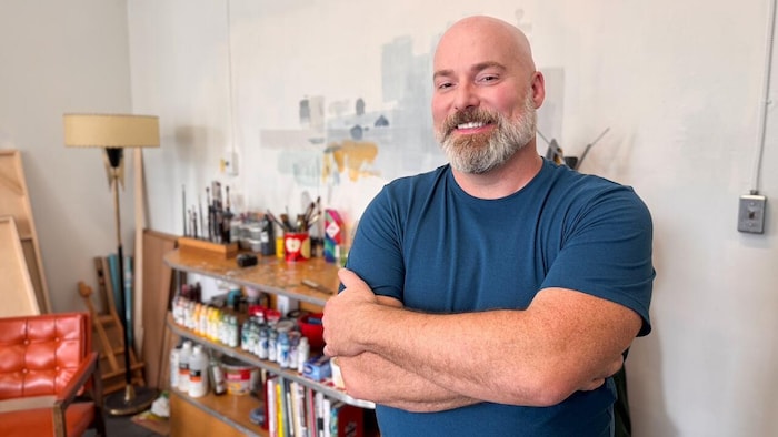Craig Guthrie sourit, les bras croisés, dans un atelier où l’on aperçoit des pinceaux et des contenants de peinture.