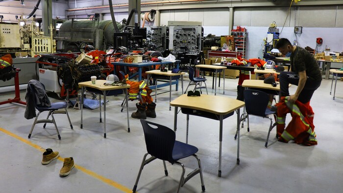 Un étudiant enfile une tenue orange de travail près de son pupitre de classe. La pièce est un immense atelier.