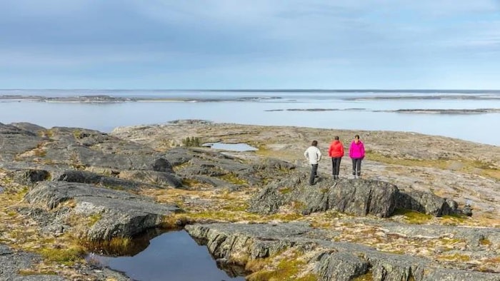 Photo de paysage. Au loin, on aperçoit trois personnes de dos sur des rochers. Elles observent la vaste étendue d'eau devant elles.  
