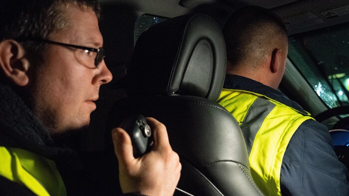 Deux hommes dans une voiture. Le passager tient un appareil de communication radio.
