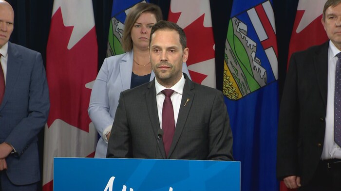 L'honorable Matt Jones, ministre des Services hospitaliers et chirurgicaux debout s'adresse à un auditoire lors d'une conférence de presse.  
