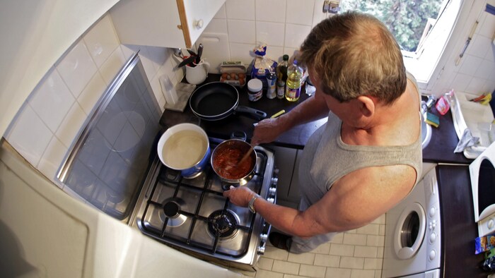 Un homme photographié de haut dans une petite cuisine. Il est penché au-dessus de la cuisinière et brasse une sauce à spaghetti dans un chaudron.