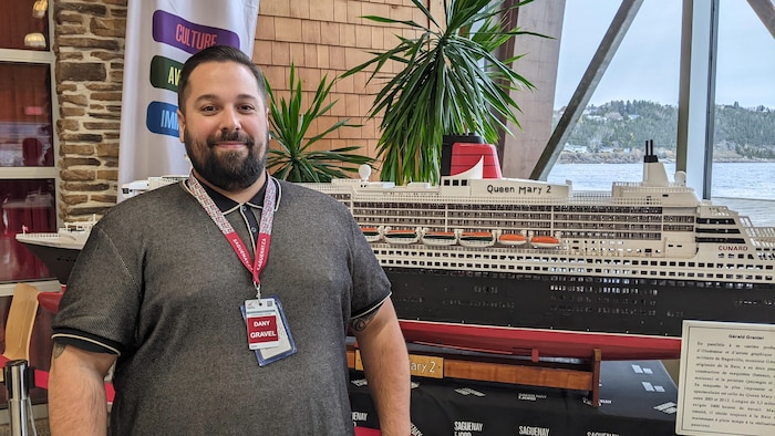 Un homme qui porte une lanière d'identification pose devant la maquette d'un bateau de croisière.