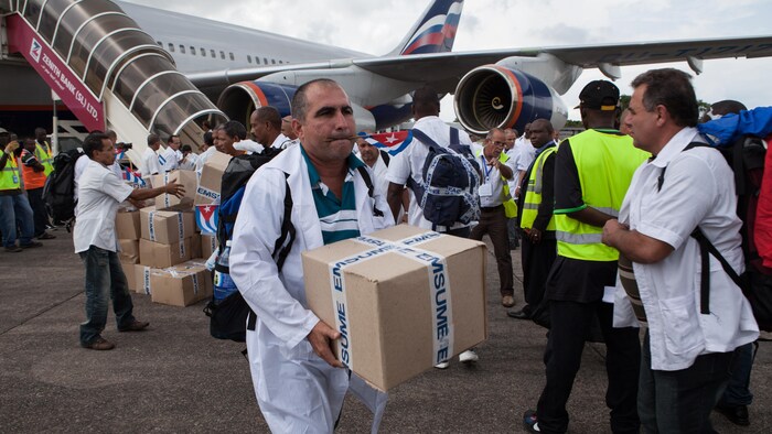 Un homme en blouse portant un carton, le drapeau de Cuba en bouche. Derrière lui, d'autres médecins qui descendent d'un avion.