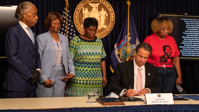 Andrew Cuomo, assis à une table et entouré de militants pour les droits des Noirs, signe un document.