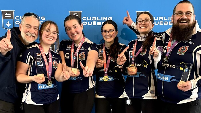 Quatre joueuses de curling et deux entraîneurs sourient à la caméra en levant l'index à titre de numéros un.